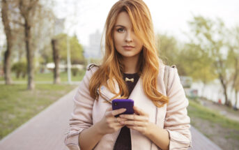 A woman with long red hair is standing on a paved path in a park, holding a purple smartphone in both hands. She is wearing a light-colored jacket and looking directly at the camera. Trees and greenery are visible in the background.