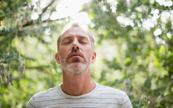 A person with short, light hair and a beard stands outdoors with eyes closed, appearing peaceful. The individual is wearing a light-colored, striped shirt and is surrounded by lush, green foliage. Soft sunlight filters through the trees in the background.