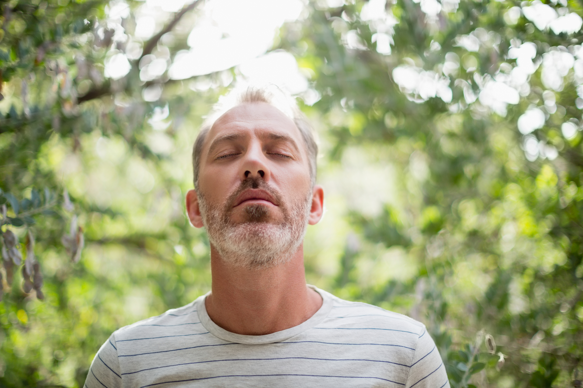 A person with short, light hair and a beard stands outdoors with eyes closed, appearing peaceful. The individual is wearing a light-colored, striped shirt and is surrounded by lush, green foliage. Soft sunlight filters through the trees in the background.