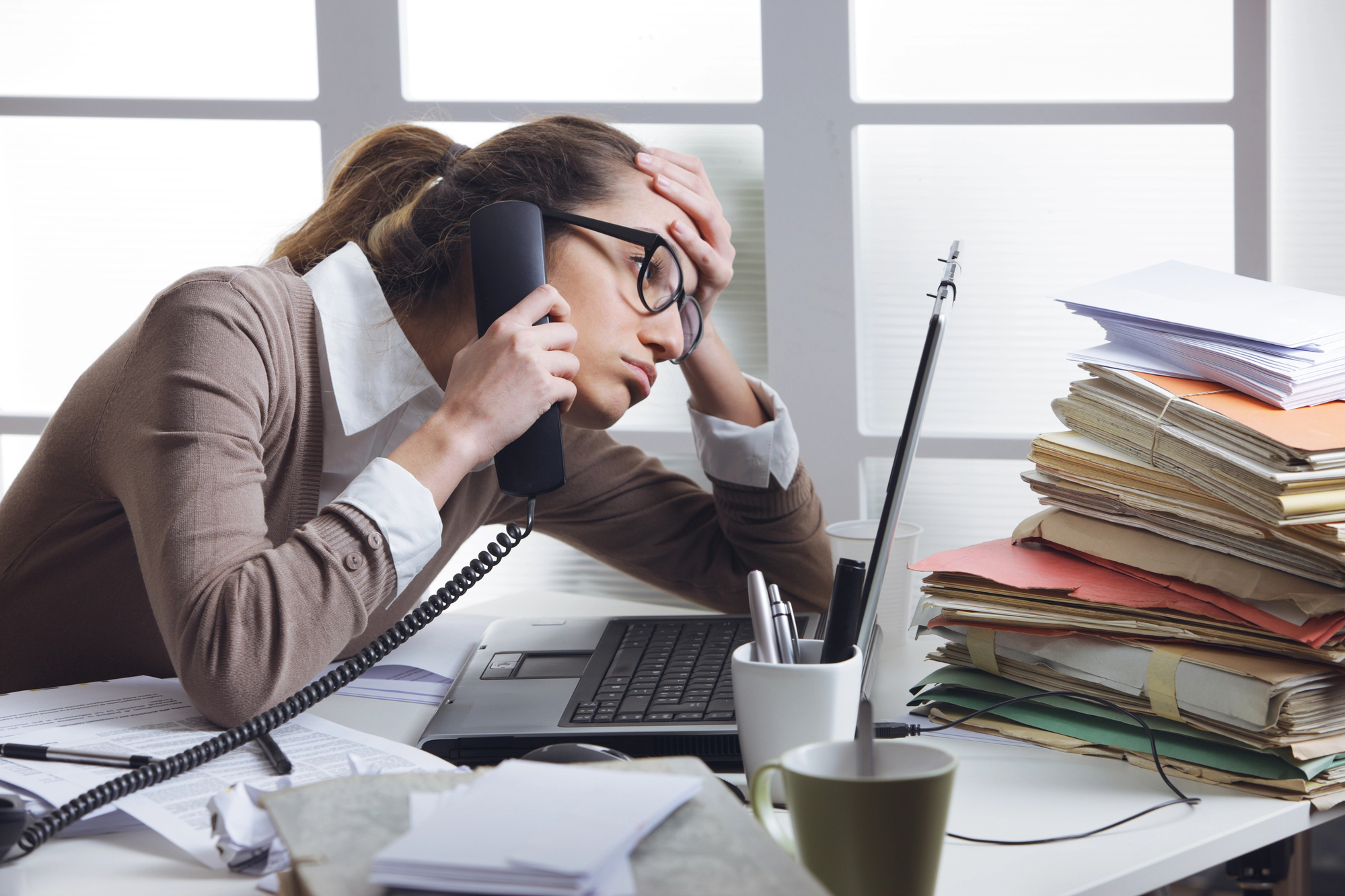 A stressed woman wearing glasses holds a phone to her ear and rests her forehead on her hand. She is seated at a cluttered desk with a laptop, stacks of papers, folders, and a cup with pens. The background features a brightly lit window.