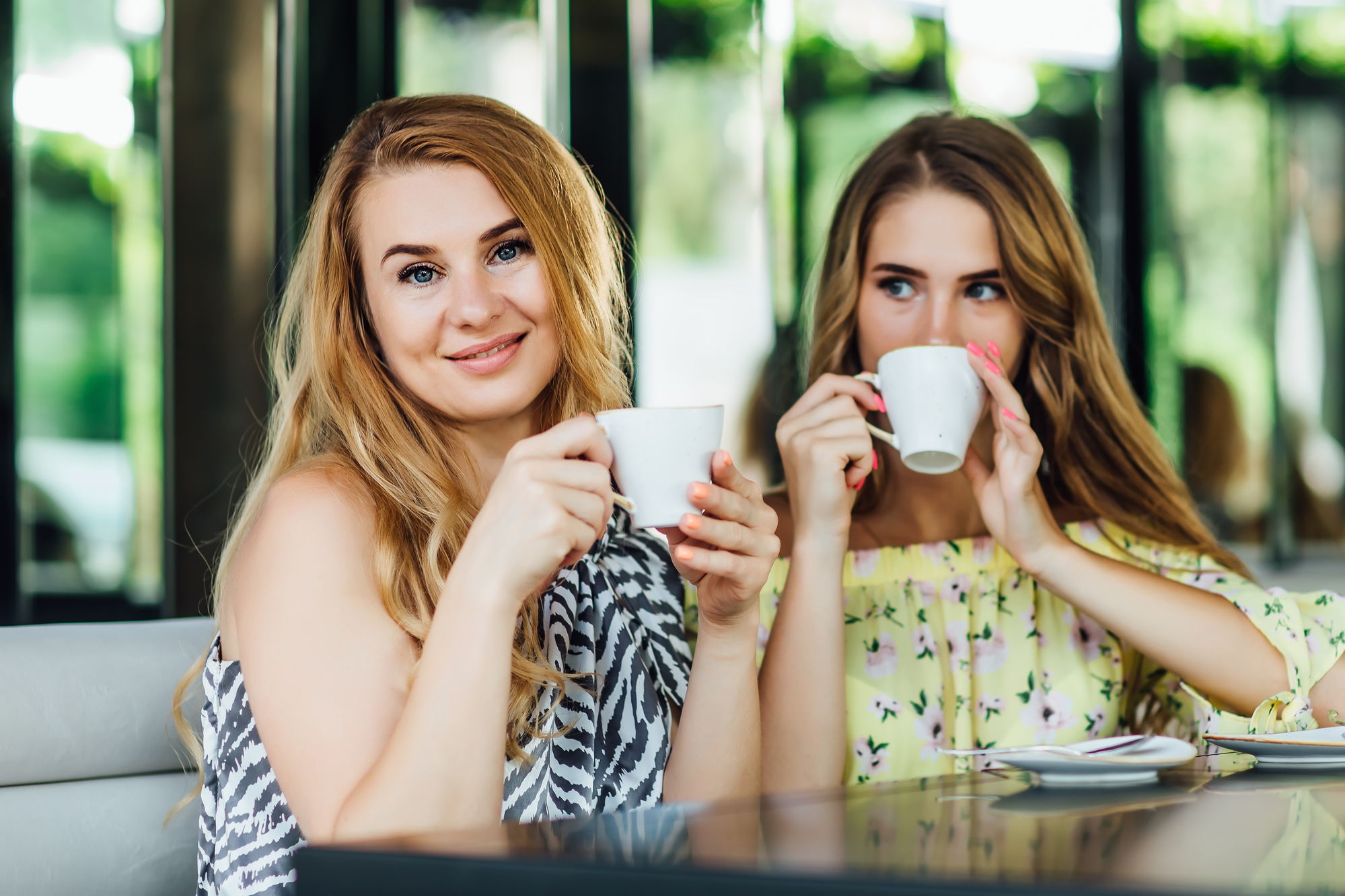 Two women sit at an outdoor café, smiling and enjoying coffee from white cups. The woman on the left has long blonde hair and wears a patterned sleeveless top, while the woman on the right has long brunette hair and wears a yellow off-shoulder top with floral designs.