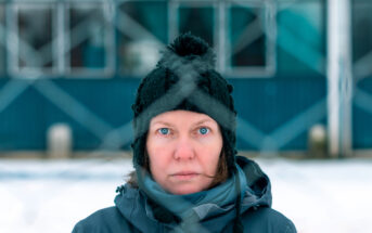 A person stands outdoors in a snowy environment, staring directly into the camera with wide blue eyes. They are wearing a dark winter hat with a pom-pom and warm clothing. In the background, a blue and white building is visible through a chain-link fence.