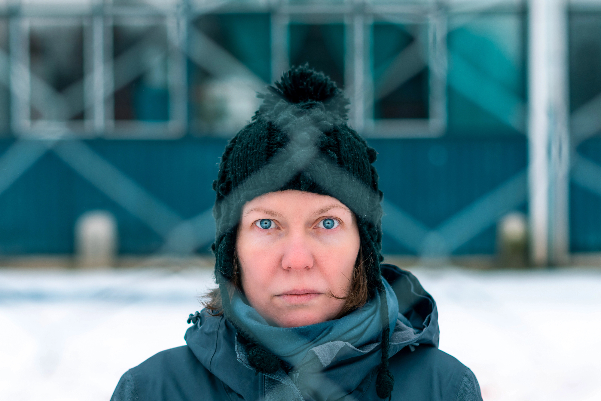 A person stands outdoors in a snowy environment, staring directly into the camera with wide blue eyes. They are wearing a dark winter hat with a pom-pom and warm clothing. In the background, a blue and white building is visible through a chain-link fence.