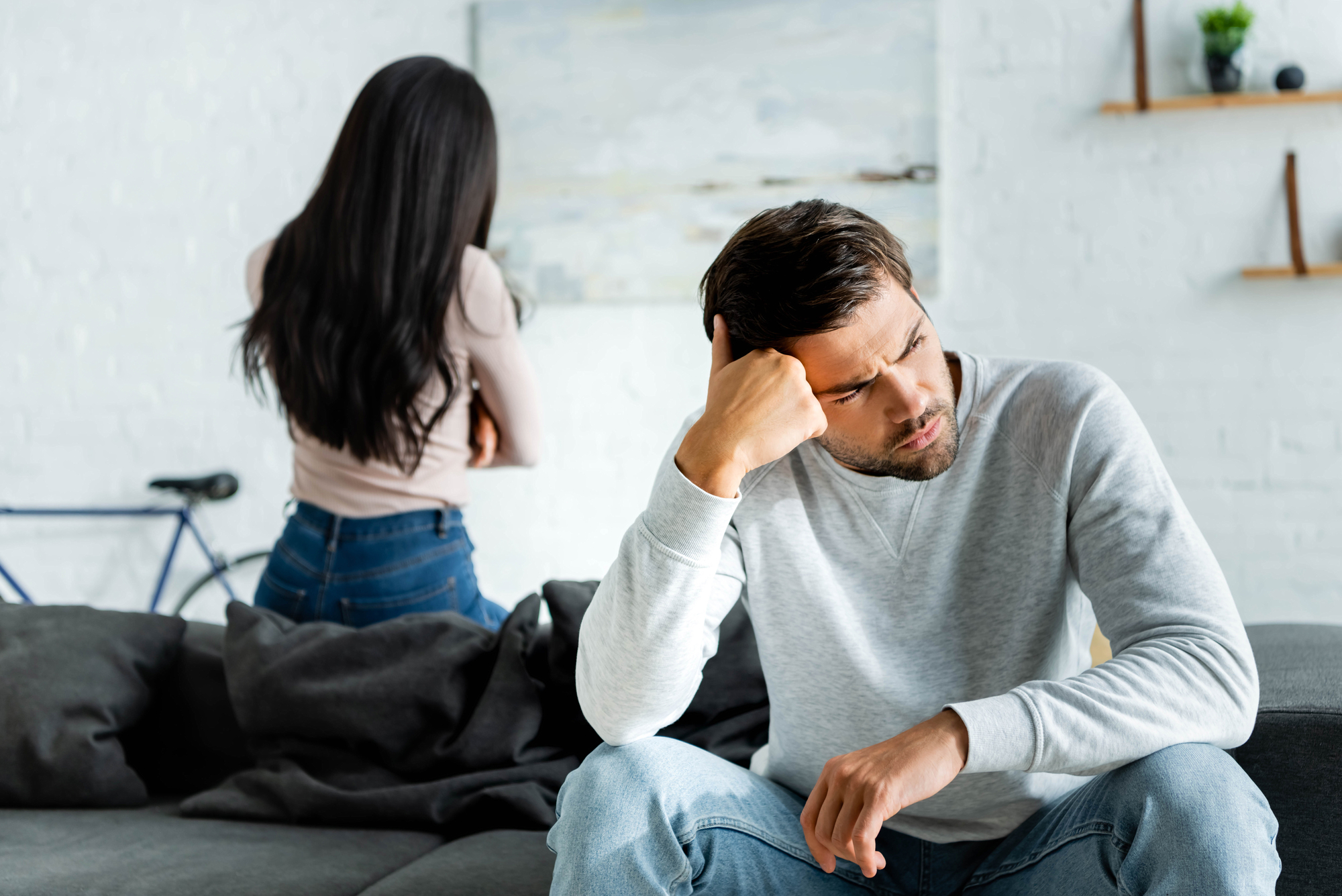 A man sitting on a couch with a distressed expression, holding his head with one hand. A woman stands in the background with her back turned to him and arms crossed. The room has a minimalist design with a white brick wall, a bicycle, and shelves.