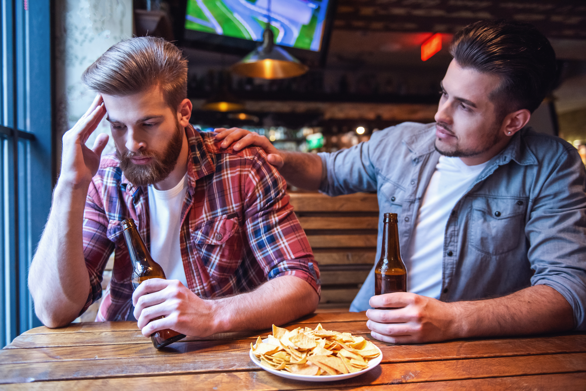 Two men are sitting at a wooden table in a bar, each holding a beer bottle. The man on the left, looking distressed, has his head resting on his hand. The man on the right is comforting him with a hand on his shoulder. A plate of nachos is on the table.