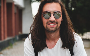 A man with long brown hair and a beard is smiling at the camera. He is wearing reflective sunglasses and a white t-shirt, with a backpack strap visible over his shoulder. The background shows a building with red bricks and a pathway.