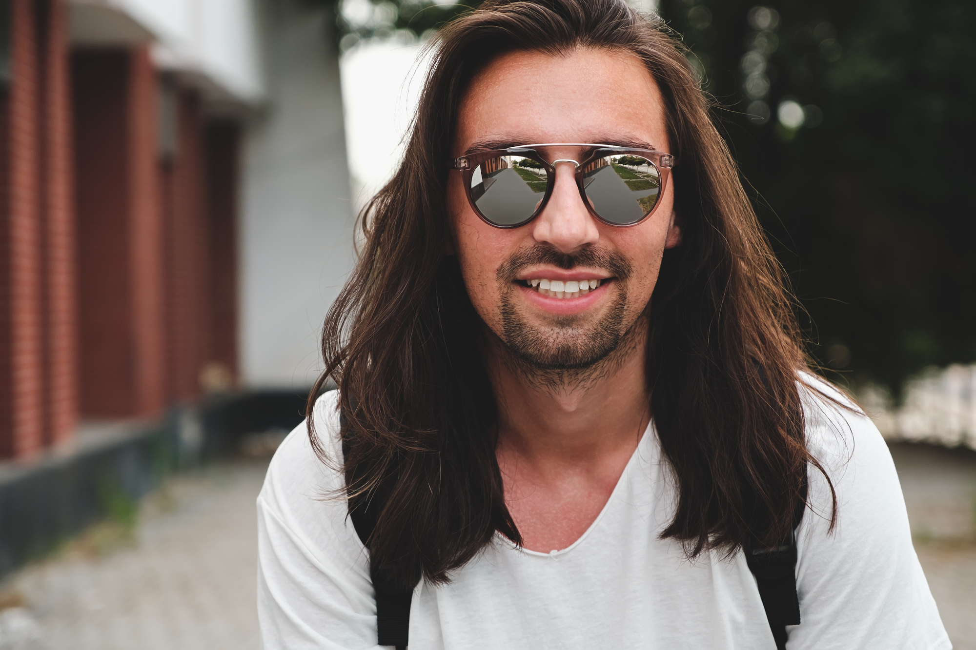 A man with long brown hair and a beard is smiling at the camera. He is wearing reflective sunglasses and a white t-shirt, with a backpack strap visible over his shoulder. The background shows a building with red bricks and a pathway.