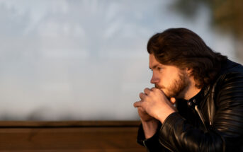 A man with medium-length hair and a beard sits pensively on a wooden bench, resting his chin on his clasped hands. He wears a black leather jacket and looks off into the distance with a thoughtful expression. The background is blurred and features soft natural light.