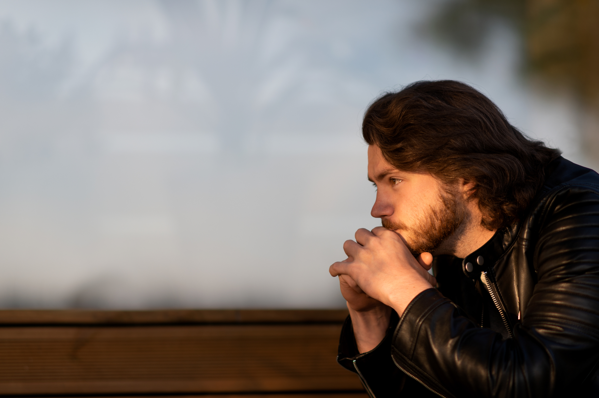 A man with medium-length hair and a beard sits pensively on a wooden bench, resting his chin on his clasped hands. He wears a black leather jacket and looks off into the distance with a thoughtful expression. The background is blurred and features soft natural light.