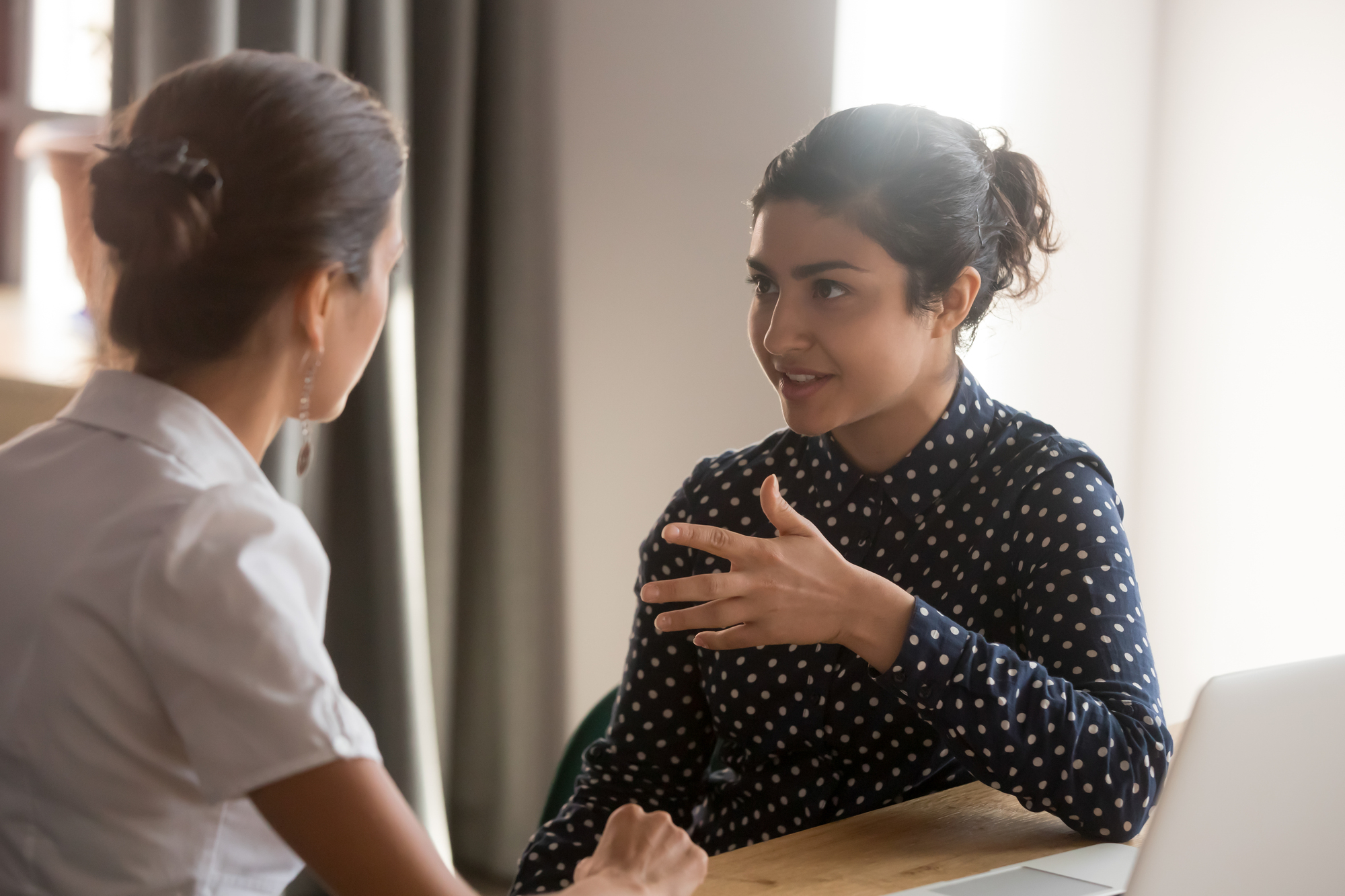 Two women are sitting at a table engaged in a conversation. The woman on the right, wearing a dark polka-dotted shirt, gestures with her hand while speaking, and the woman on the left, wearing light-colored clothing, listens attentively. A laptop is on the table.