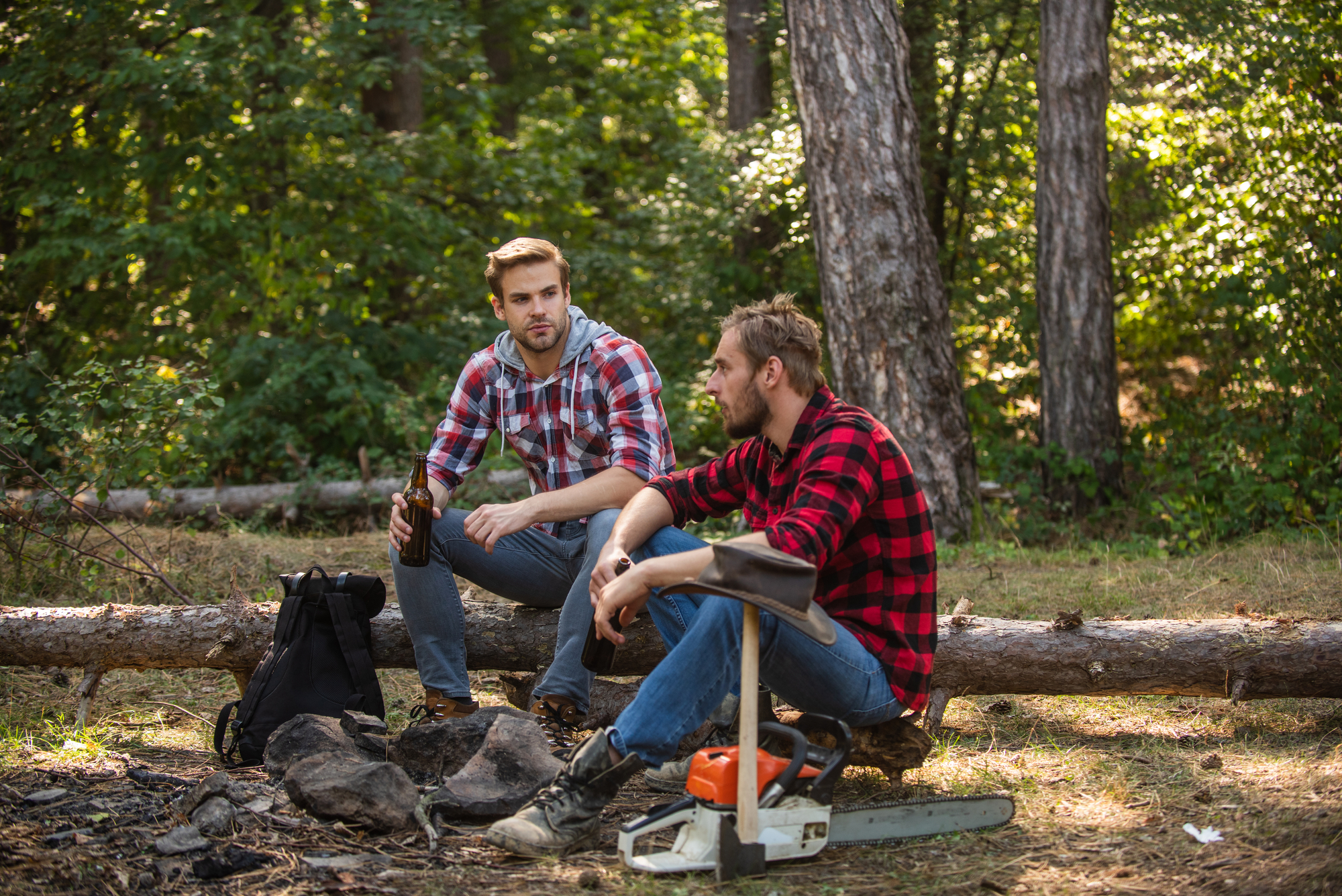 Two men in plaid shirts are sitting on logs in a forest clearing. One is holding a bottle while the other, next to a chainsaw, has his arms resting on his knees. A backpack is on the ground near them. Tall trees and green foliage surround the scene.