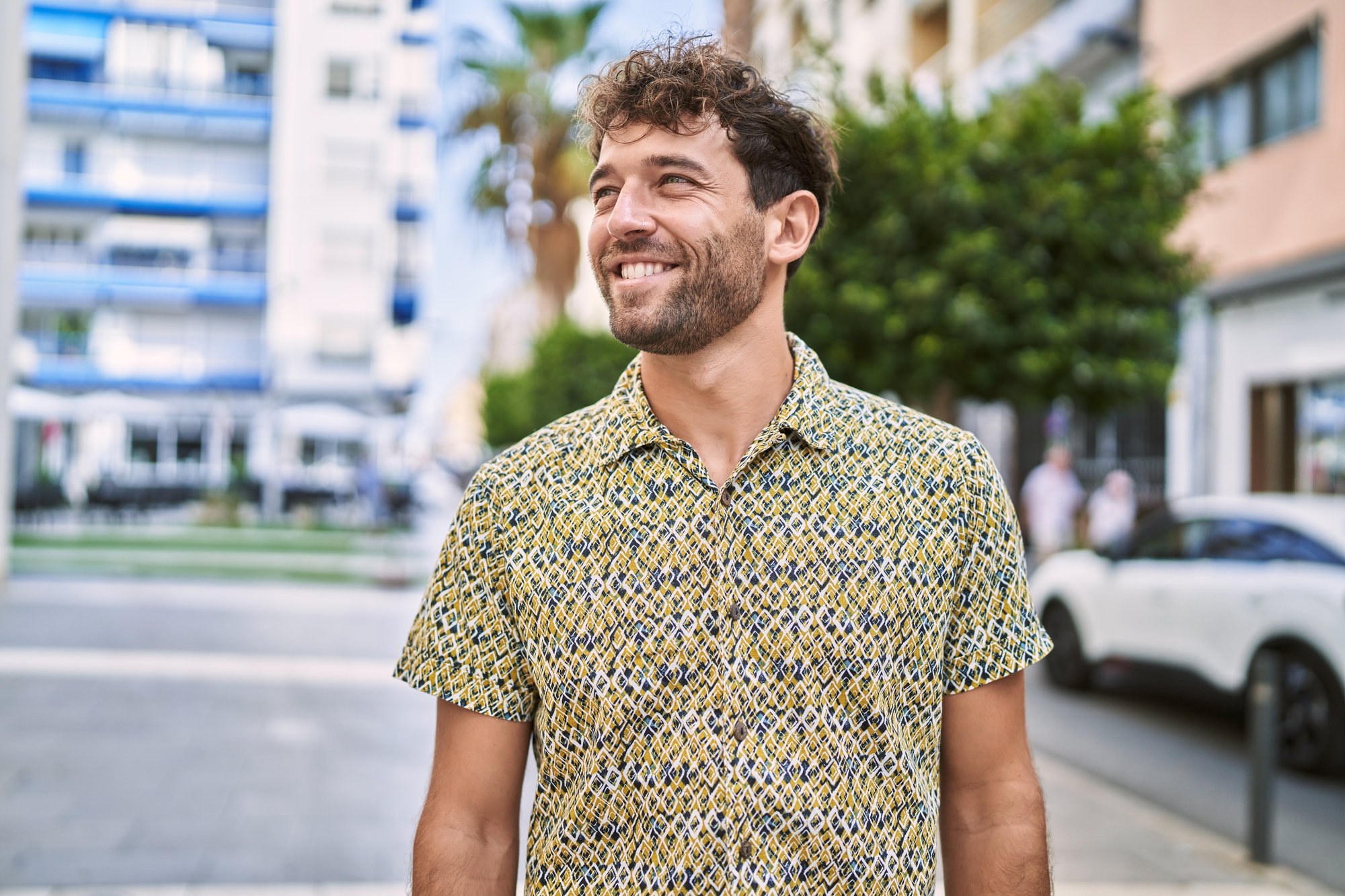 A man with curly hair and a beard is smiling while standing outdoors on a sunny day. He is wearing a patterned short-sleeved shirt. Trees, buildings, and a parked white car are visible in the background.