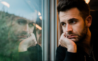 A man with short dark hair and a beard leans against a train window, resting his chin on his hand and looking out. His reflection is visible in the glass, showing a thoughtful or pensive expression. The blurred scenery outside the window suggests the train is moving.