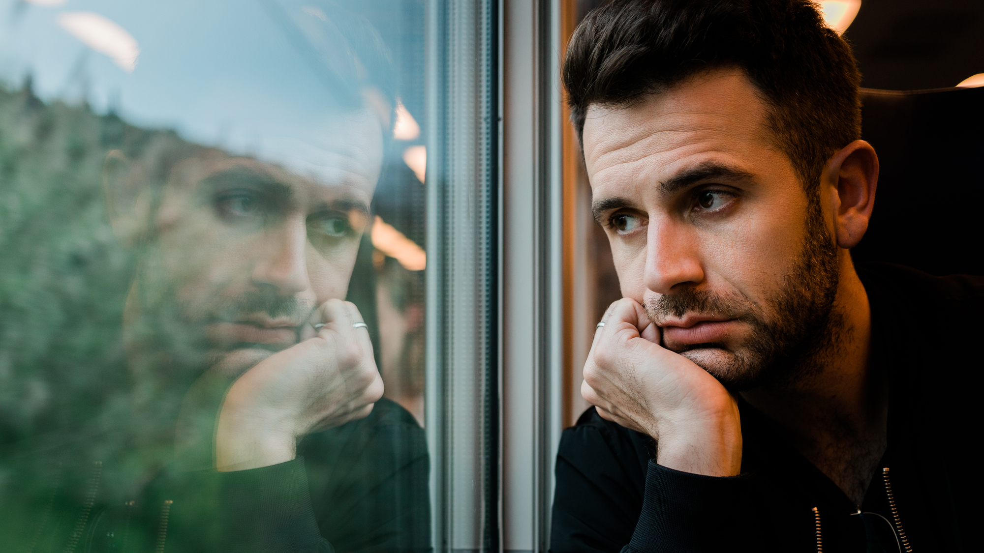 A man with short dark hair and a beard leans against a train window, resting his chin on his hand and looking out. His reflection is visible in the glass, showing a thoughtful or pensive expression. The blurred scenery outside the window suggests the train is moving.