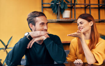 A man and a woman sit at a table, engaging in a lively conversation. The man, with a watch on his wrist, listens attentively with his chin resting on his hands, while the woman, wearing a yellow sweater, gestures animatedly. The background is a warm, mustard-yellow wall with shelves and plants.