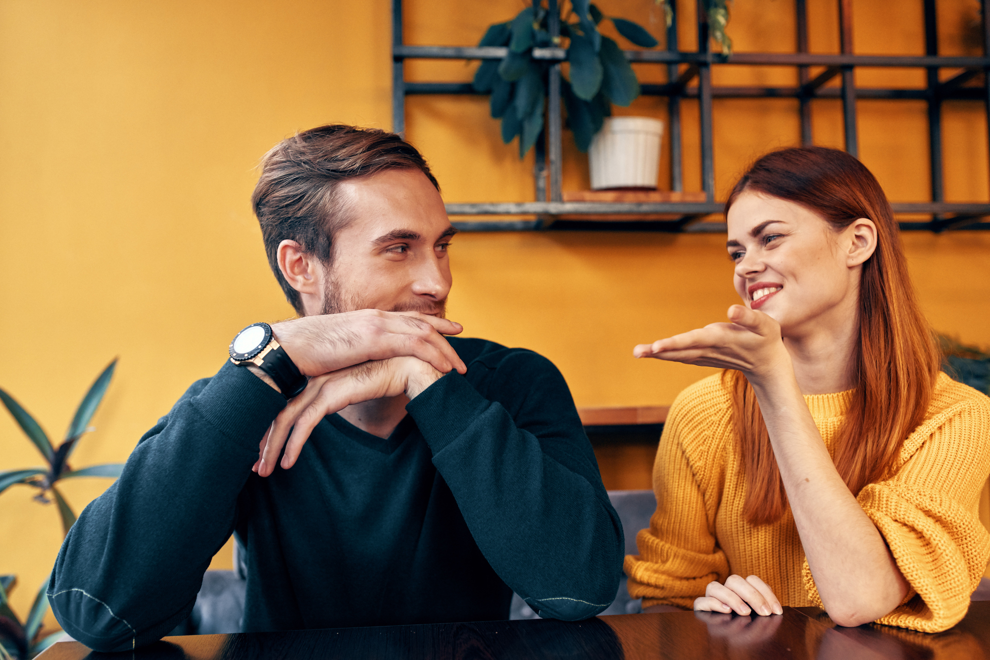 A man and a woman sit at a table, engaging in a lively conversation. The man, with a watch on his wrist, listens attentively with his chin resting on his hands, while the woman, wearing a yellow sweater, gestures animatedly. The background is a warm, mustard-yellow wall with shelves and plants.