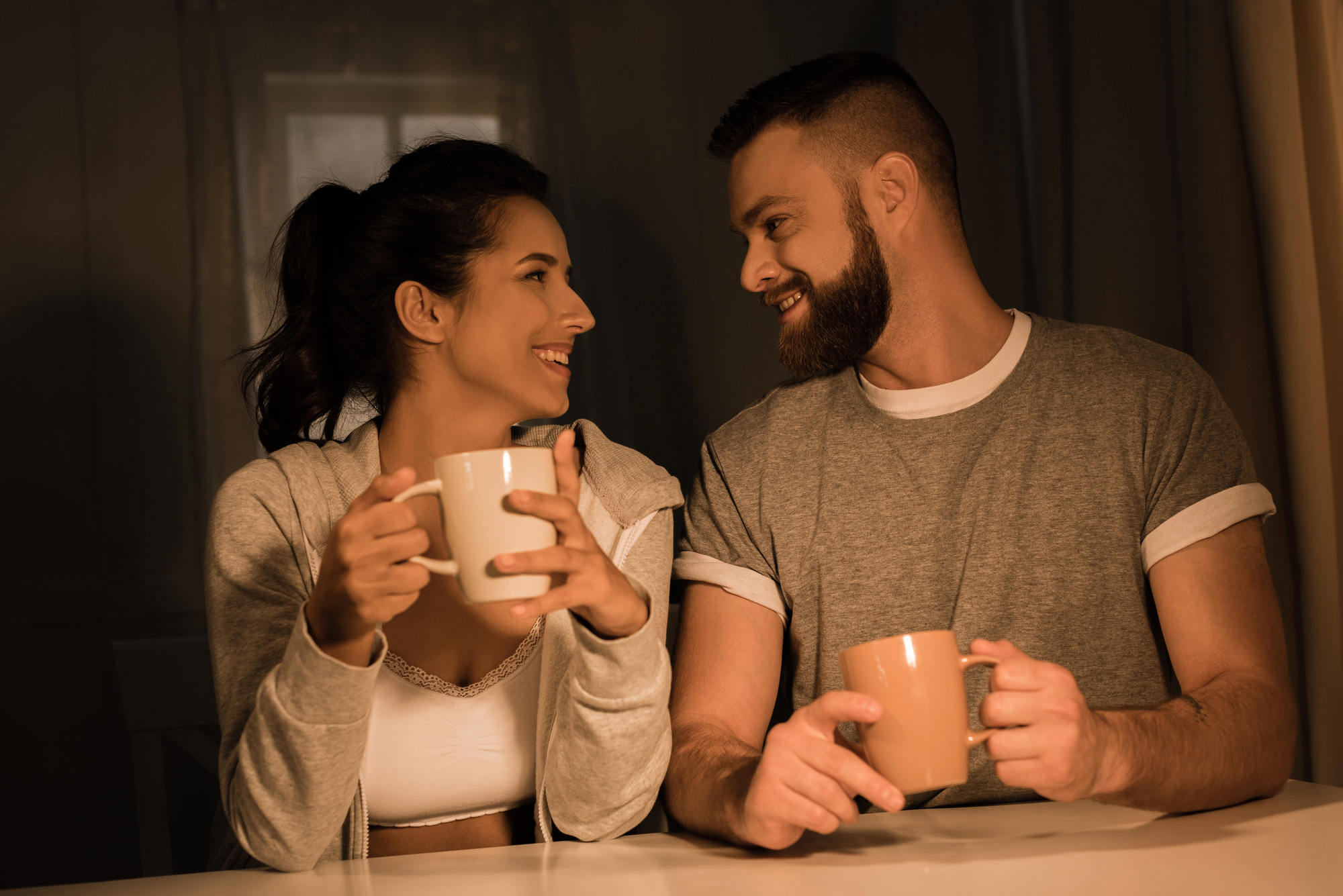 A couple sits closely together at a table, smiling at each other warmly. They are both holding beige mugs, with a cozy, relaxed atmosphere created by soft lighting. The woman has her hair in a ponytail, and the man has a beard and short hair.