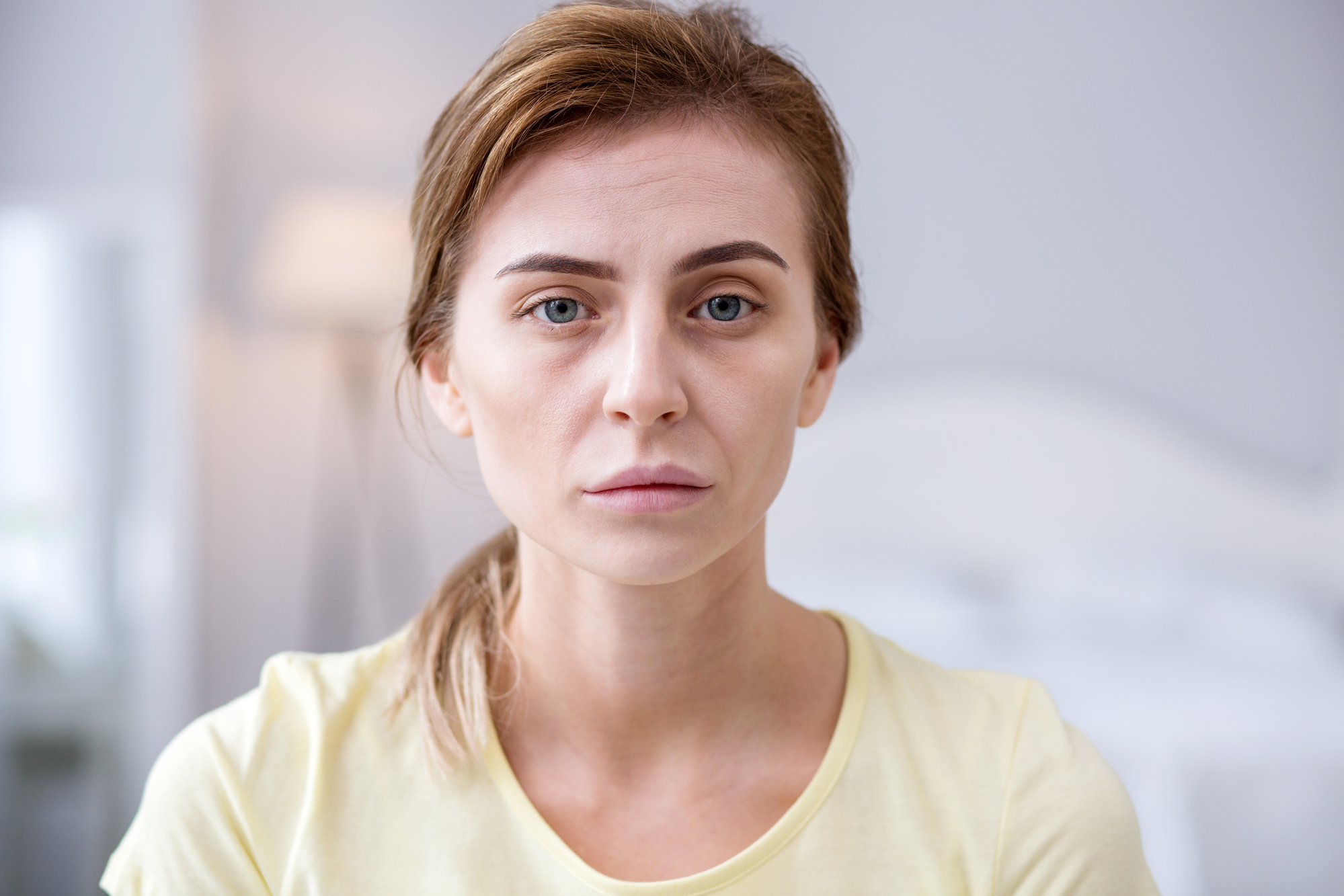 A woman with light brown hair pulled back in a ponytail is looking directly at the camera with a neutral expression. She is wearing a light yellow shirt. The background is softly blurred, showing a bed and a lamp, suggesting an indoor setting.