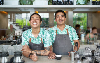 Two baristas standing behind a counter in a bright, modern café. Both wearing matching green tropical print shirts and grey aprons, one is smiling broadly with arms resting on the counter, while the other holds a coffee cup, also smiling. Various coffee tools are visible.