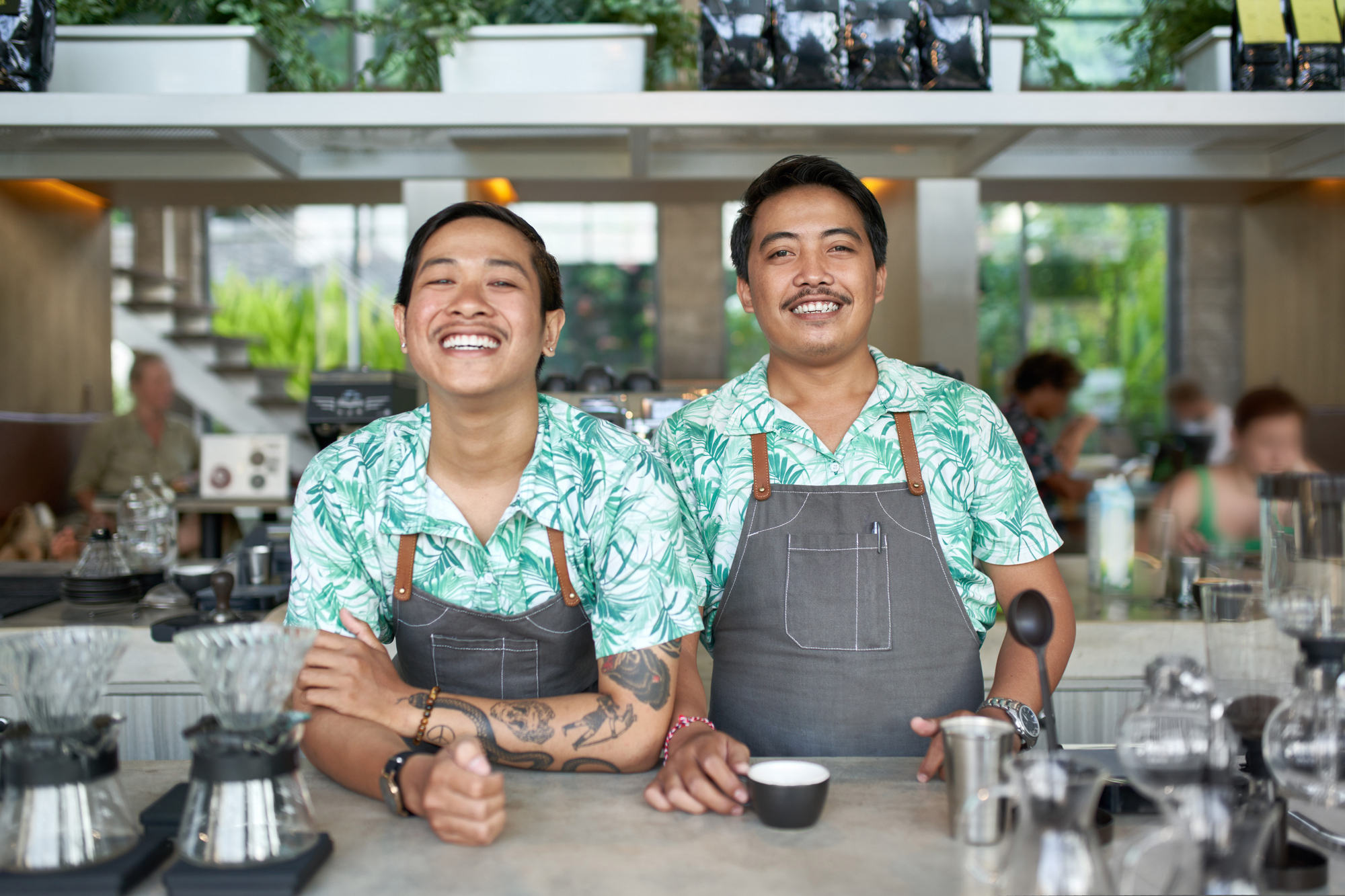 Two baristas standing behind a counter in a bright, modern café. Both wearing matching green tropical print shirts and grey aprons, one is smiling broadly with arms resting on the counter, while the other holds a coffee cup, also smiling. Various coffee tools are visible.