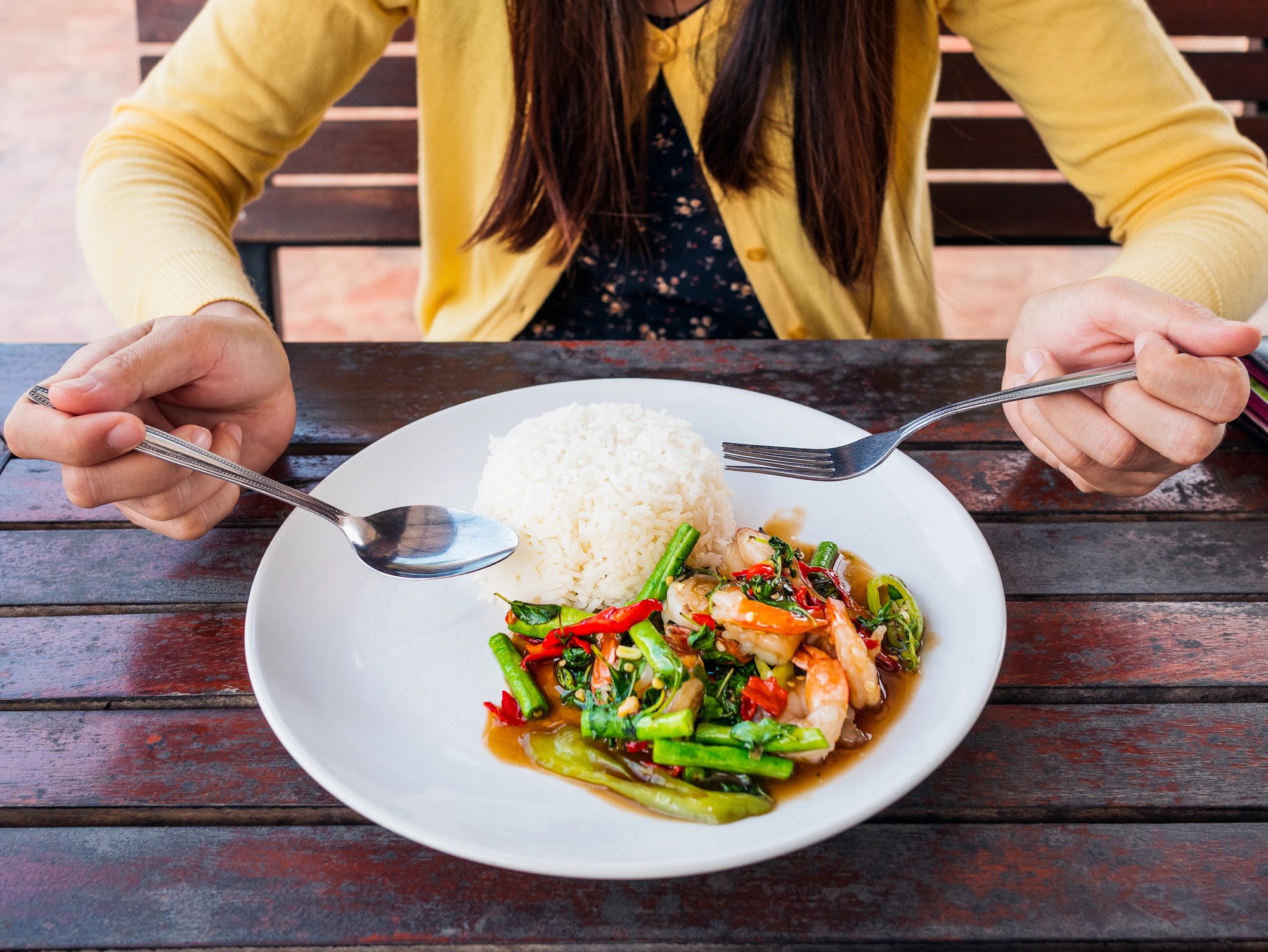 A person in a yellow cardigan sitting at a wooden table with a plate of food. The plate contains a serving of white rice and a colorful stir-fry dish with vegetables and shrimp. The person is holding a fork and a spoon, ready to eat.