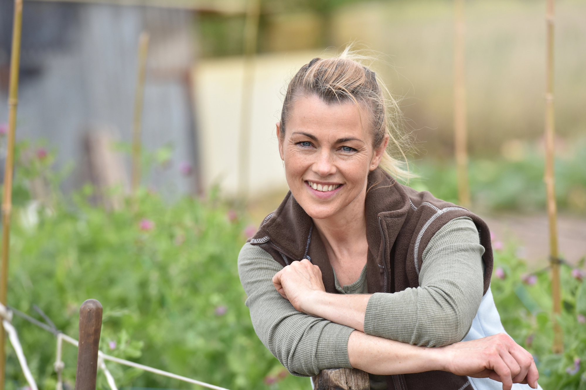 A woman with blond hair tied back smiles while leaning on a wooden fence in a garden. She is wearing a green long-sleeve shirt and a brown vest. Blurry green plants and garden structures are visible in the background.