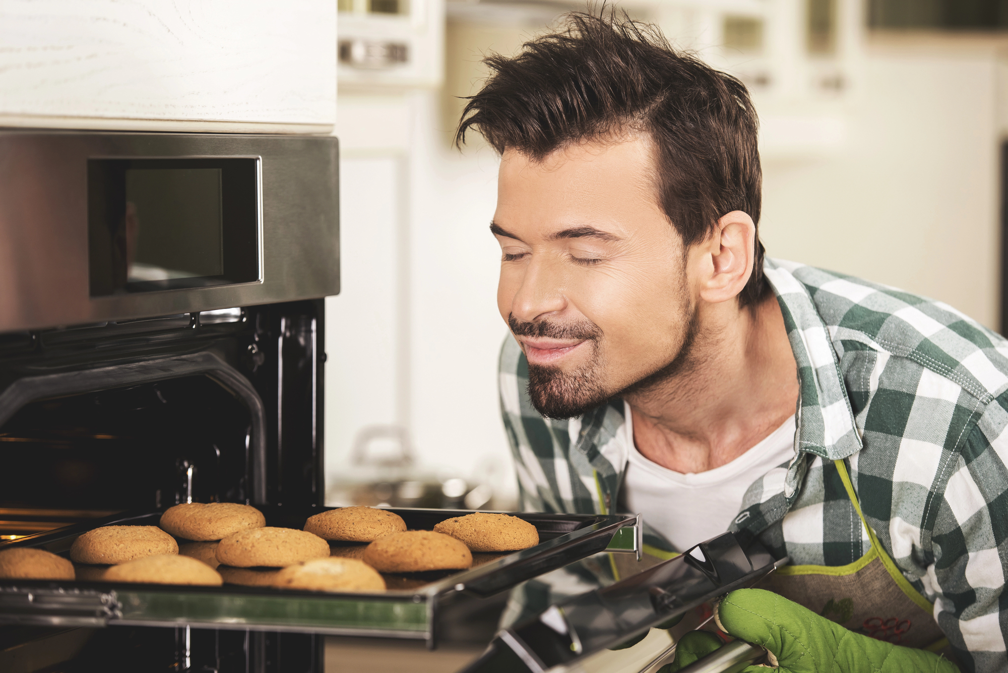 A man with short hair, wearing a plaid shirt and an apron, smiles contentedly as he leans toward an open oven, holding a tray of freshly baked cookies. The kitchen in the background has white cabinets and counters.