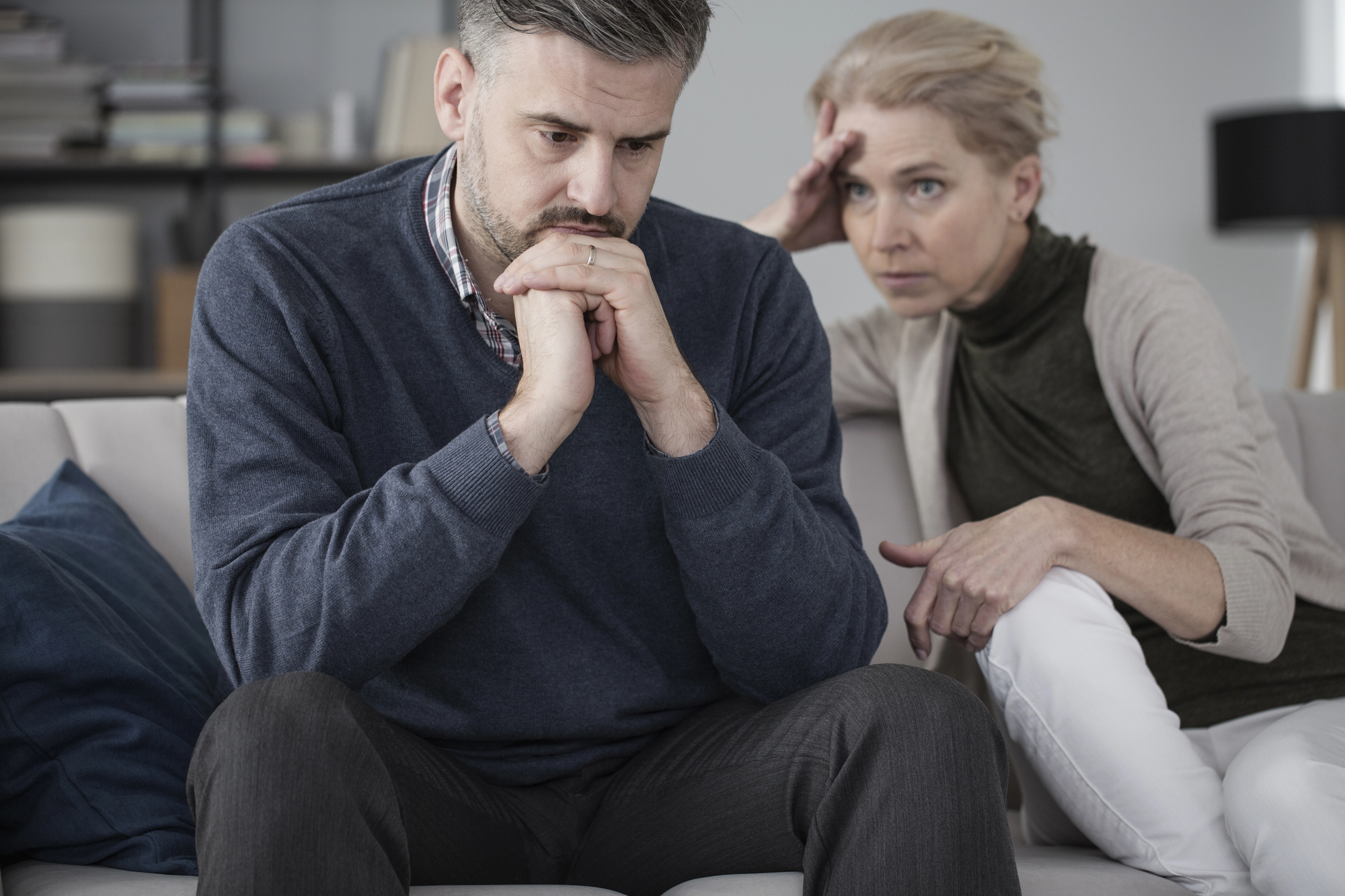 A man and woman sit on a couch, both looking concerned. The man, wearing a blue sweater, leans forward with his hands clasped under his chin. The woman, in a green top, sits beside him with one hand on her forehead, appearing worried. Shelves are visible in the background.