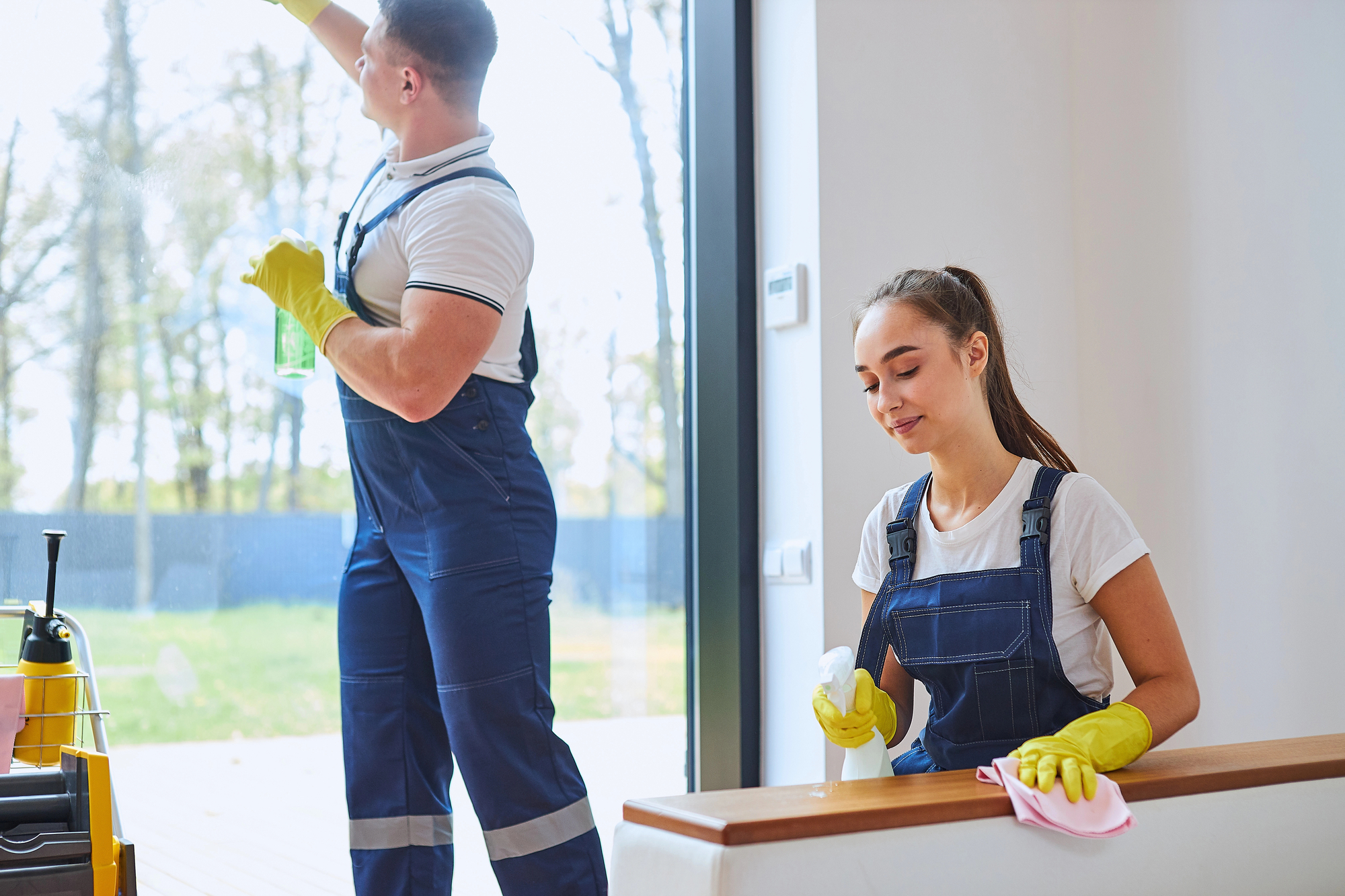 Two workers in blue overalls and yellow gloves are cleaning. The woman in the foreground is wiping a wooden surface with a cloth, while the man in the background is spraying and cleaning a large window. A cleaning cart is partially visible.