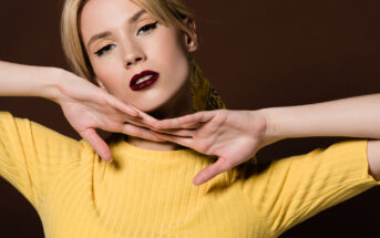 A woman with blonde hair styled in a loose updo poses against a brown background. She wears deep red lipstick and a yellow top, with her hands elegantly framing her face. She gazes confidently at the camera.