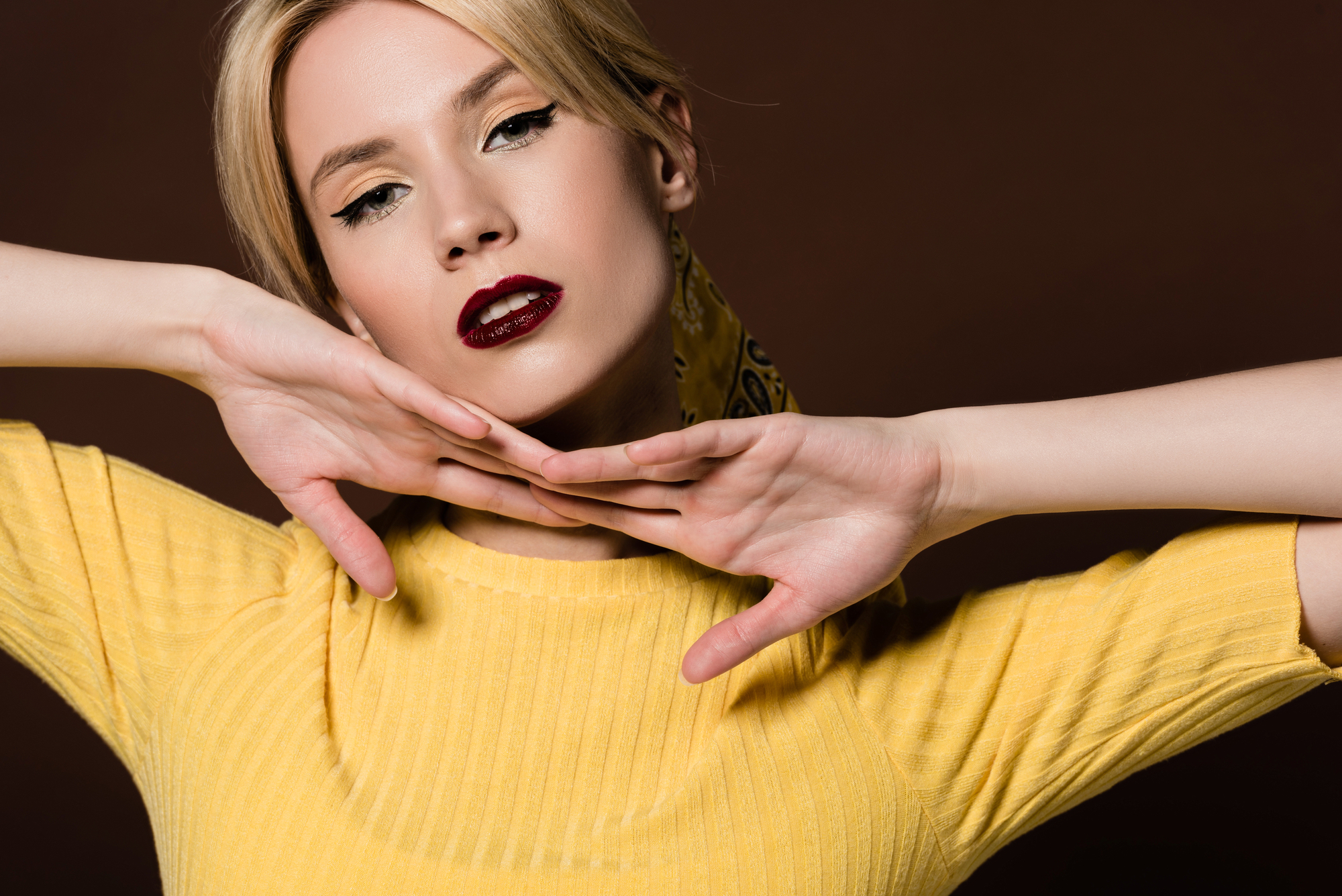 A woman with blonde hair styled in a loose updo poses against a brown background. She wears deep red lipstick and a yellow top, with her hands elegantly framing her face. She gazes confidently at the camera.