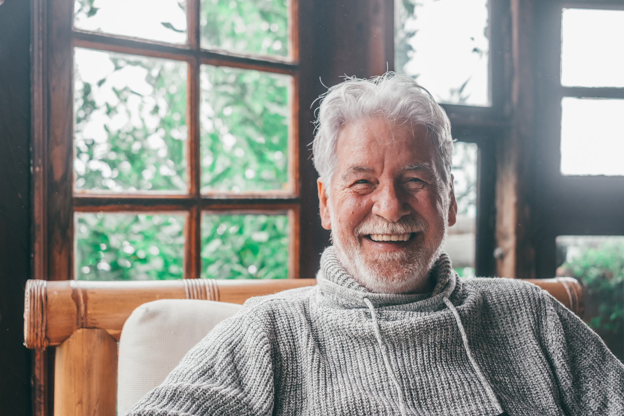 An older man with white hair and a beard is sitting indoors on a light-colored couch, wearing a gray knit sweater. He is smiling broadly, with large windows showing greenery behind him. The room appears cozy and warmly lit.