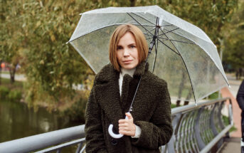 A woman with short blonde hair, wearing a dark green textured coat, stands on a bridge holding a clear umbrella. The background features trees and a body of water, suggesting an overcast or rainy day.