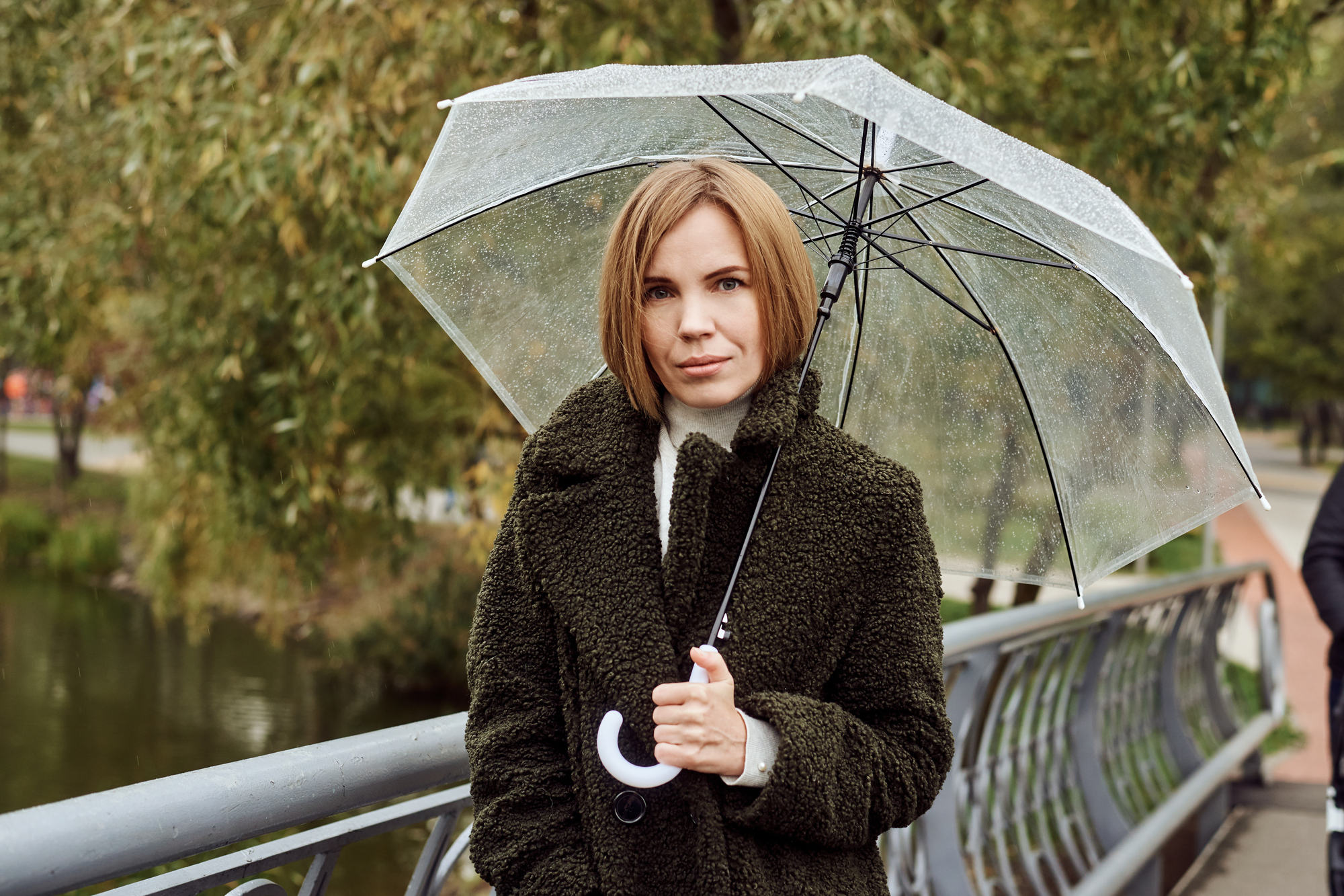 A woman with short blonde hair, wearing a dark green textured coat, stands on a bridge holding a clear umbrella. The background features trees and a body of water, suggesting an overcast or rainy day.