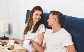 A man and a woman sit up in bed, facing each other and smiling. Both are holding white mugs. A breakfast tray with pancakes, blueberries, and a small bowl is placed on the bed between them. The bed has blue pillows, and a lamp is on the nightstand.