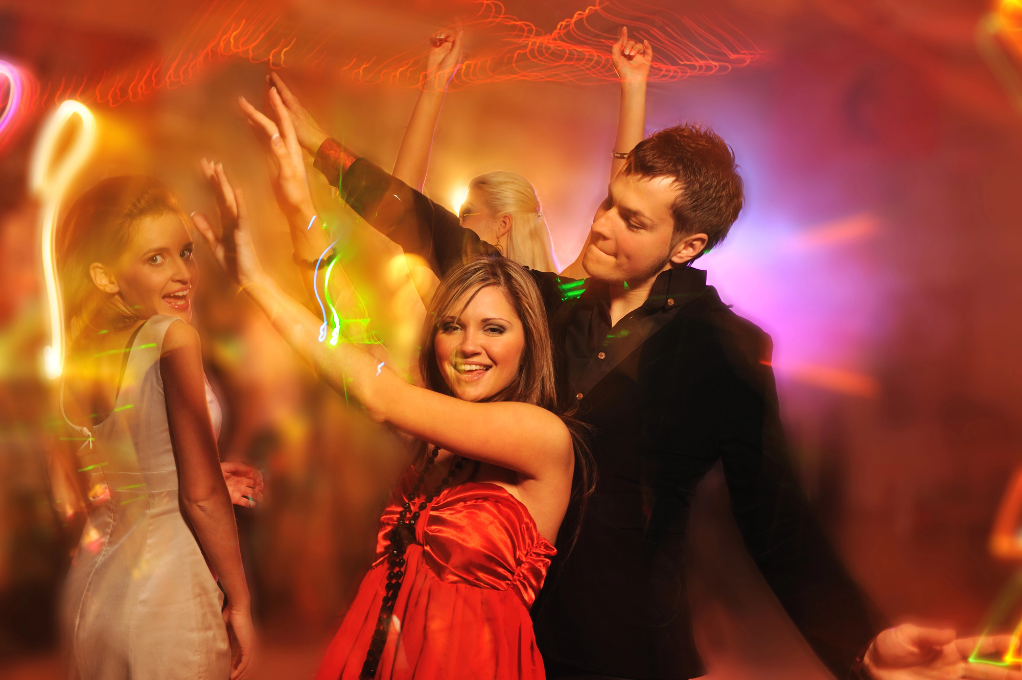 Group of young adults dancing energetically in a dimly lit, colorful room with vibrant light trails. A woman in a red dress and a man in a black shirt are in the foreground, smiling and raising their arms. The atmosphere is lively and festive.