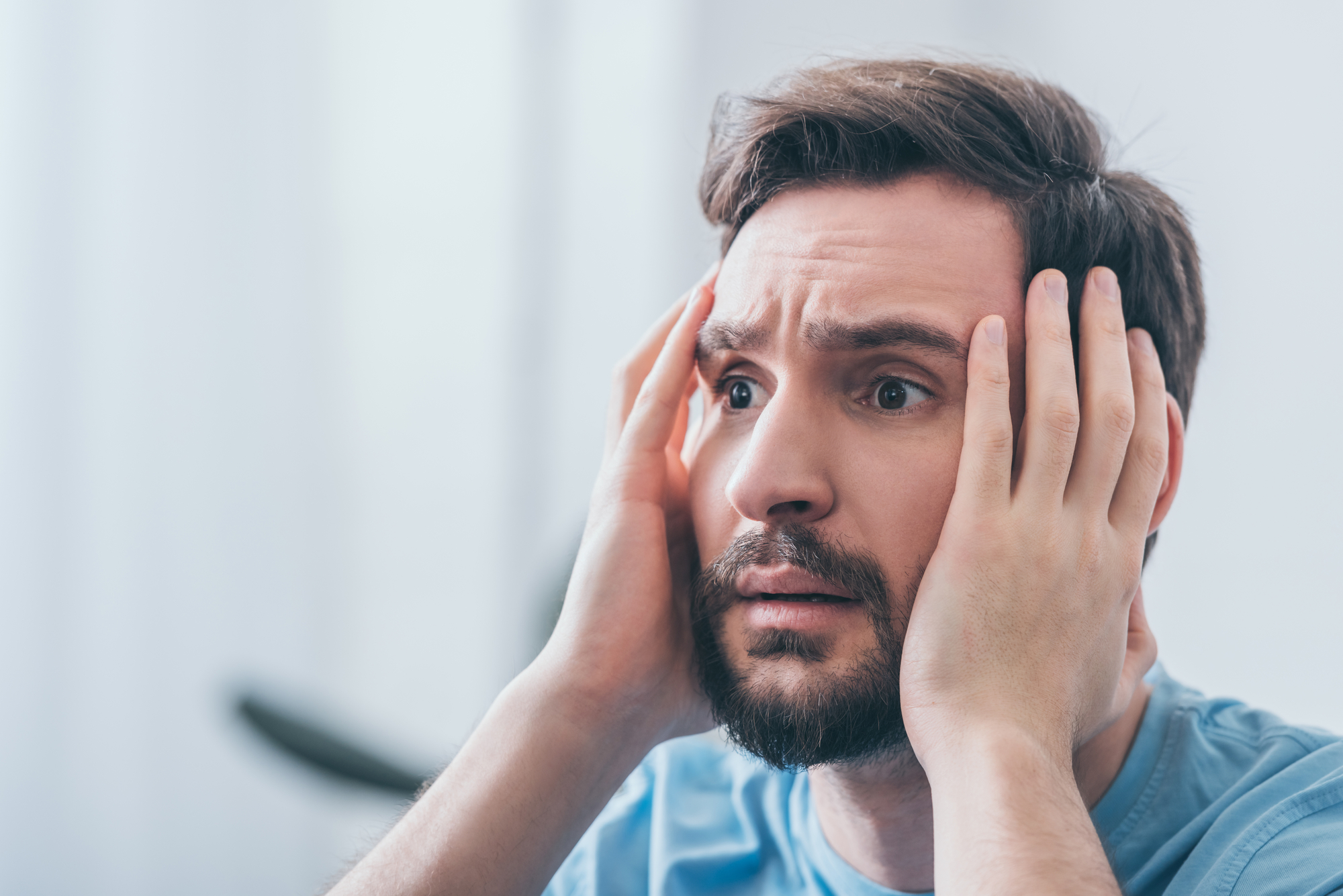 A man with a beard and short hair is holding his face with both hands, looking distressed or worried. He is wearing a light blue shirt and has a pensive expression. The background is blurred and light-colored.