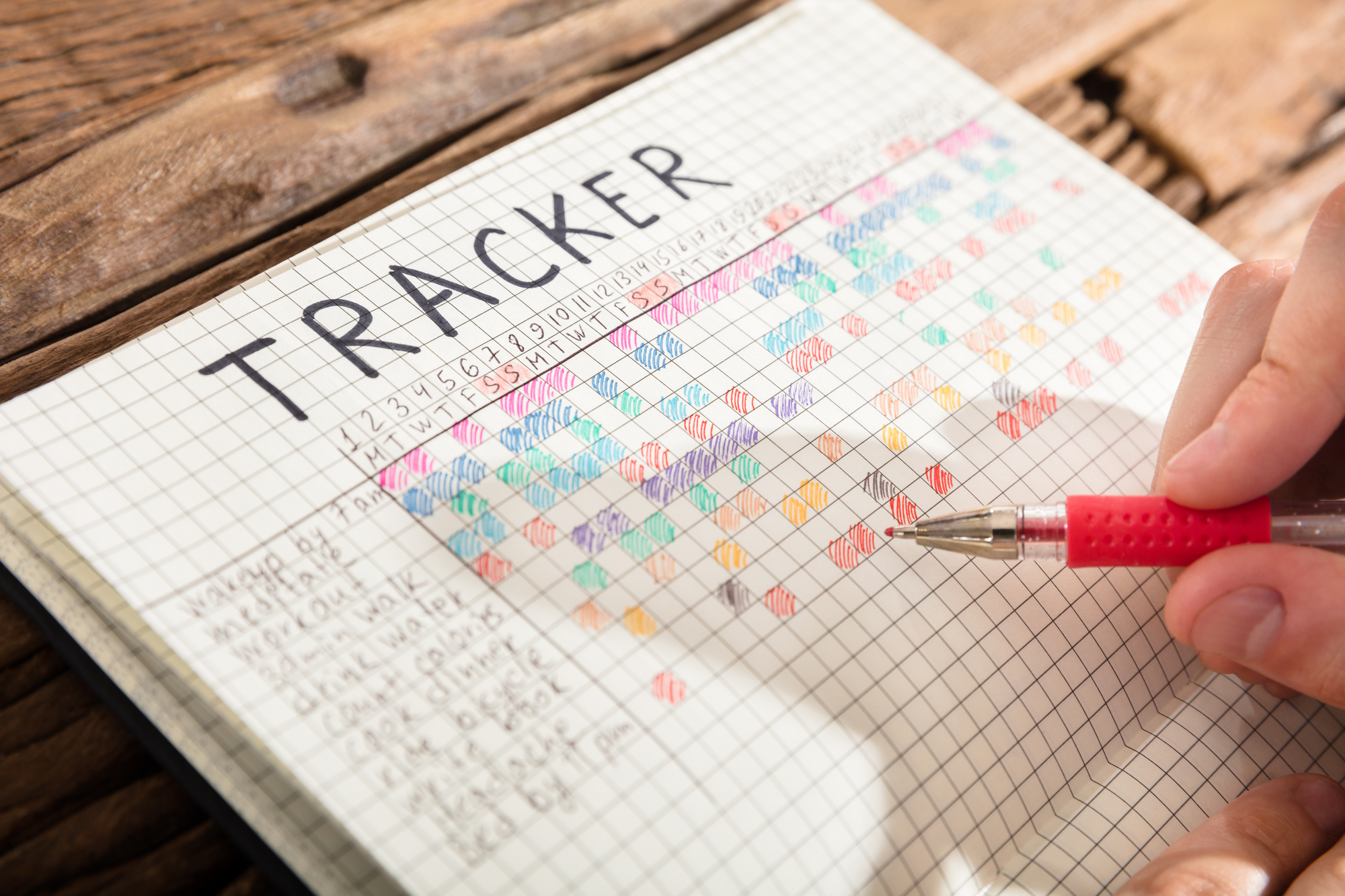 A hand holding a red pen marks a colorful tracker grid in a notebook. The notebook displays the word "TRACKER" at the top and has multiple columns and rows filled with different colored squares, indicating tracked activities or habits. The notebook rests on a wooden surface.