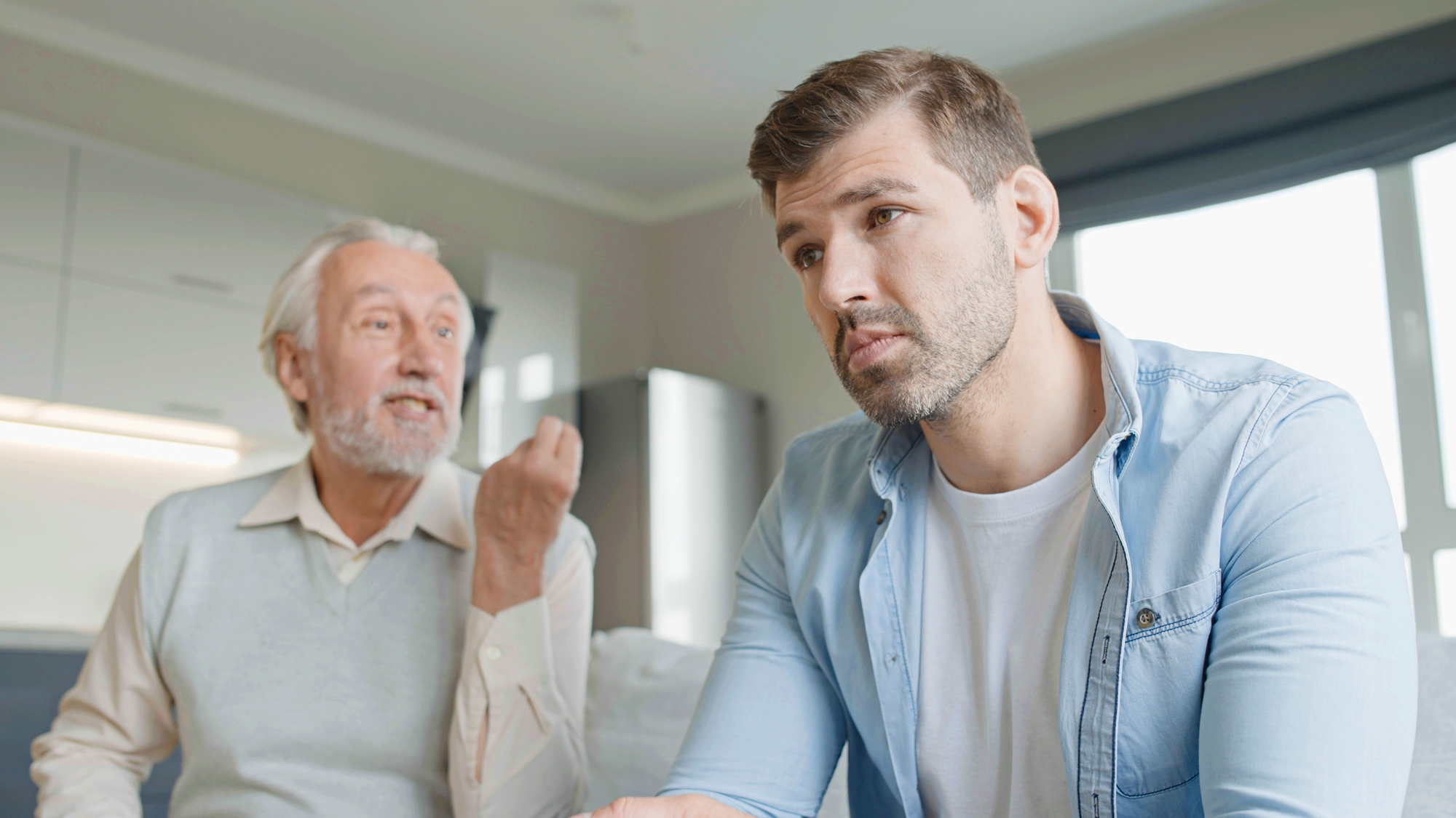 An older man with white hair and a beard passionately gestures with his hand while speaking to a younger, bearded man in a blue shirt who looks frustrated. They are sitting in a modern, brightly lit kitchen.