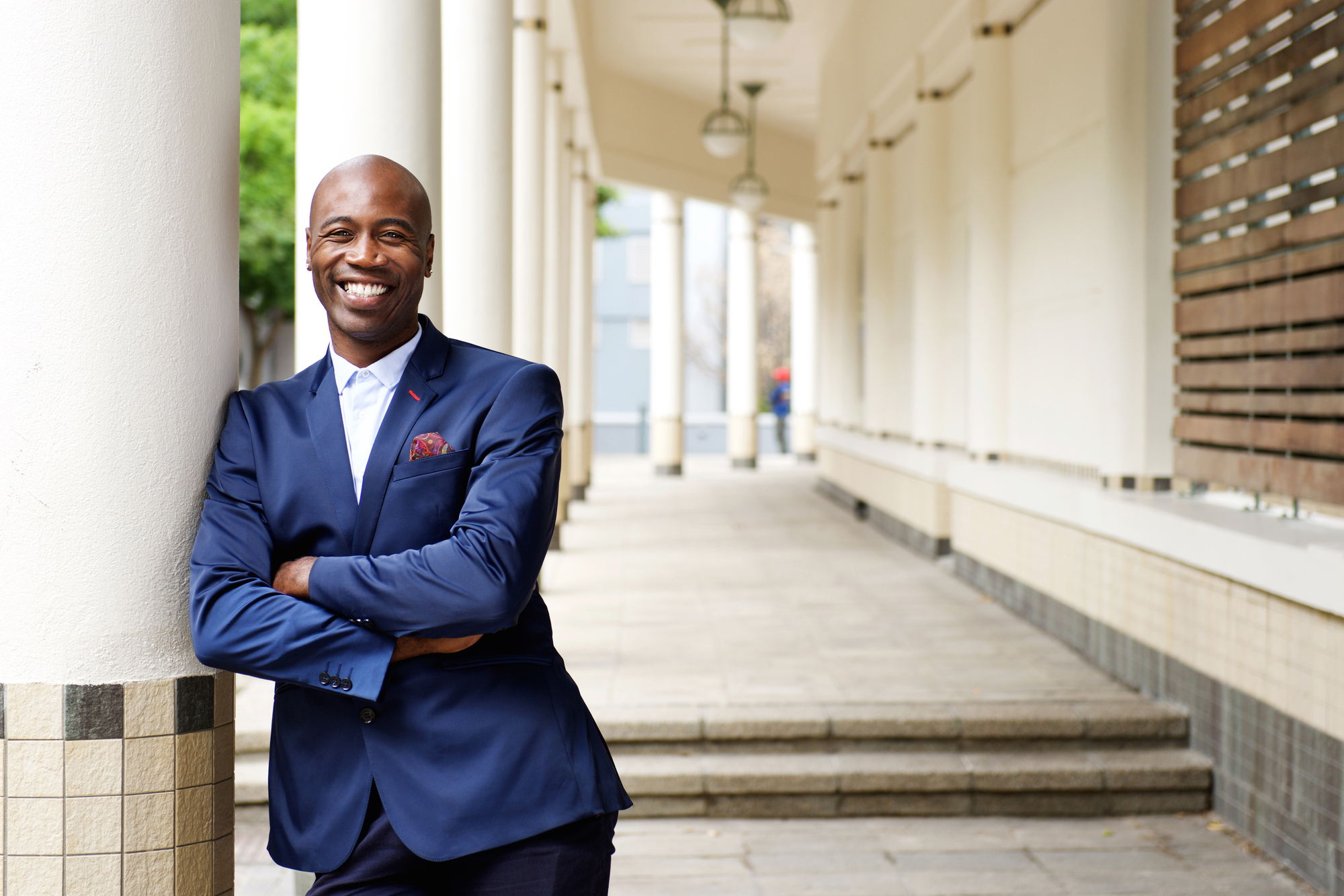 A man in a dark blue suit leans against a column with his arms crossed, smiling at the camera. He stands in a long, covered walkway with tall columns. The background includes greenery and blurred details of the building's structure.