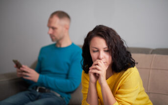 A woman with dark hair and a yellow sweater sits on a couch, looking thoughtful and concerned with her hands near her face. In the blurred background, a man in a blue sweater sits, focused on his phone, creating a sense of tension between them.