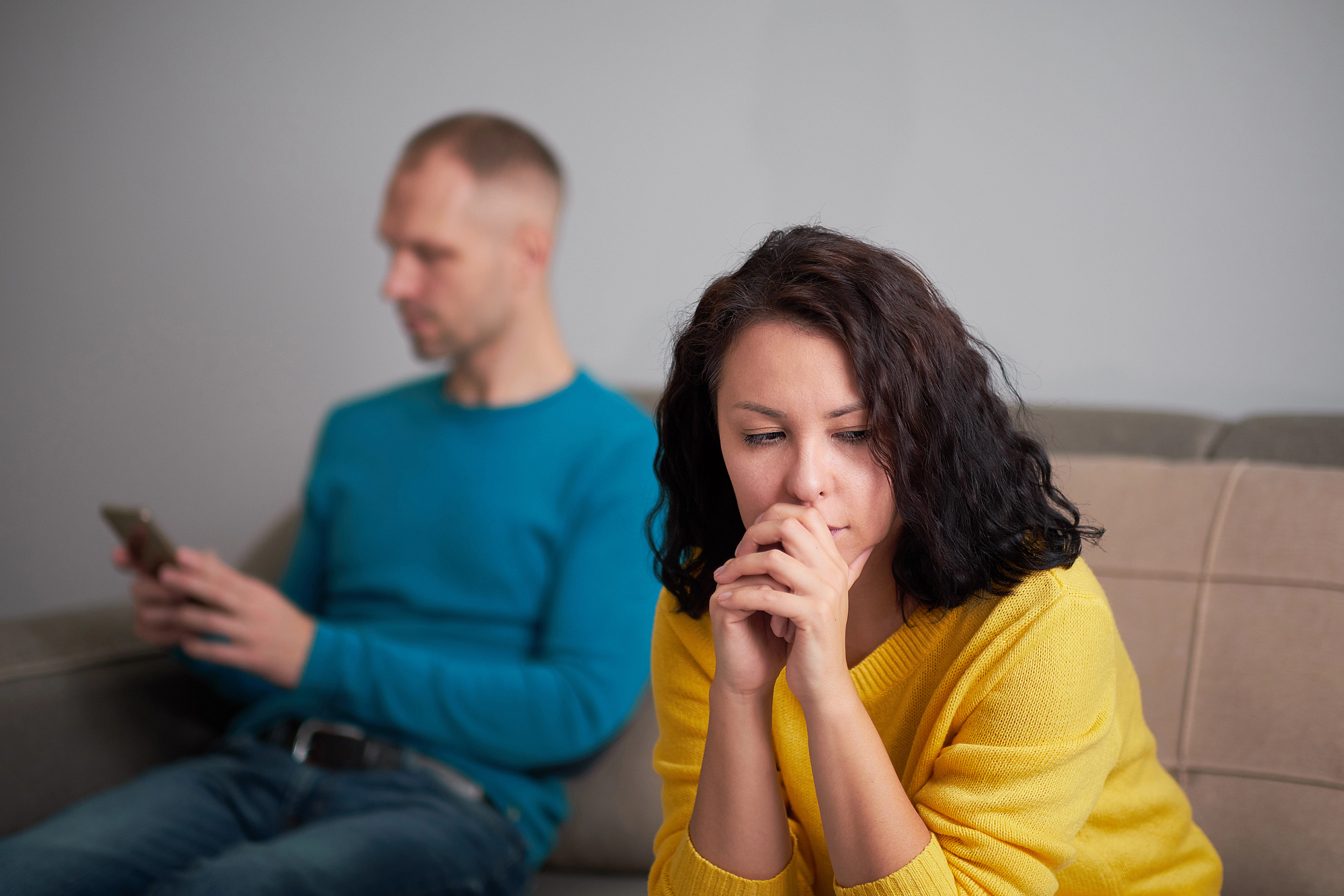 A woman with dark hair and a yellow sweater sits on a couch, looking thoughtful and concerned with her hands near her face. In the blurred background, a man in a blue sweater sits, focused on his phone, creating a sense of tension between them.