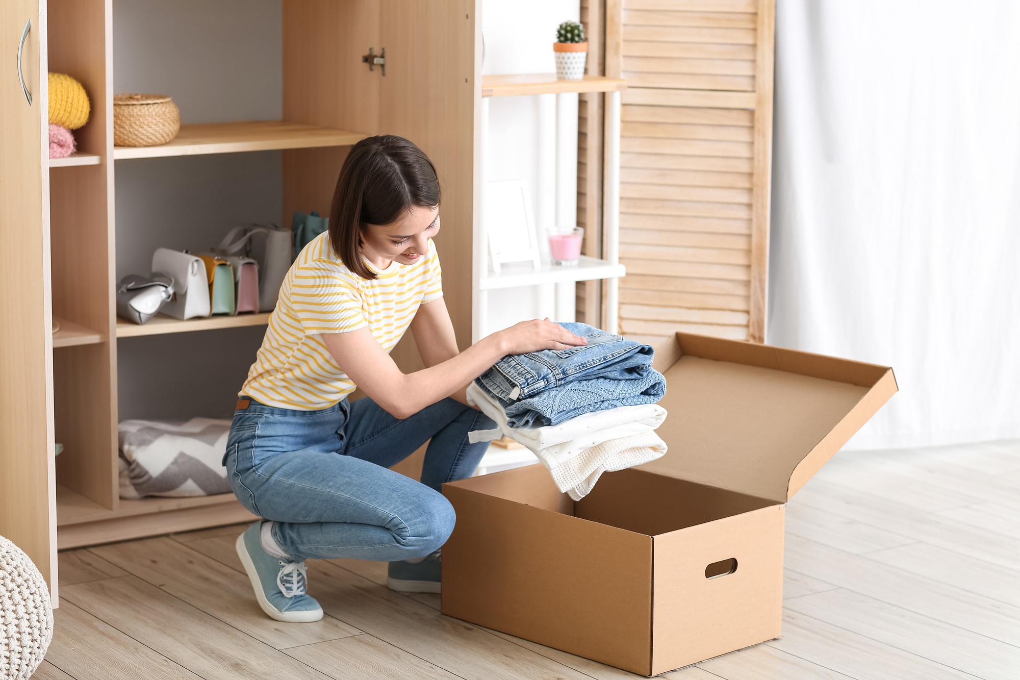 A young woman wearing a yellow striped shirt and jeans is kneeling on the floor, carefully placing folded clothes into an open cardboard box. Behind her, a wardrobe with shelves containing various household items is partially visible.