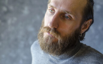 A man with a beard and mustache, wearing a grey turtleneck sweater, is gazing contemplatively away from the camera. He has light-colored eyes, an earring in his left ear, and is set against a mottled grey background.