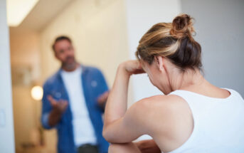 A woman with a bun leans her head on her hand, looking distressed. A man in a blue shirt stands in front of her, gesturing with his hands as if explaining or arguing. The atmosphere appears tense. The background is indoors with neutral colors.