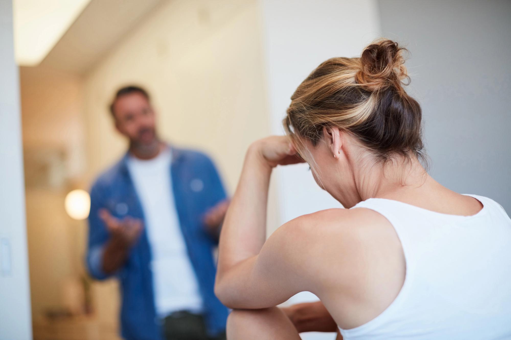 A woman with a bun leans her head on her hand, looking distressed. A man in a blue shirt stands in front of her, gesturing with his hands as if explaining or arguing. The atmosphere appears tense. The background is indoors with neutral colors.