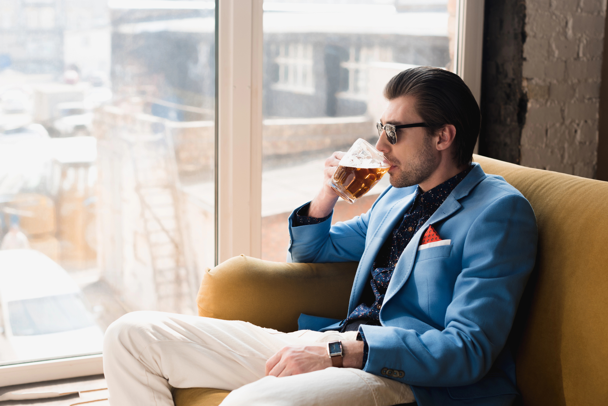 A man in a blue blazer and sunglasses is sitting on a yellow couch, sipping a drink from a glass. He has dark hair slicked back and is wearing light-colored pants. The background shows a bright window with an urban exterior scene.