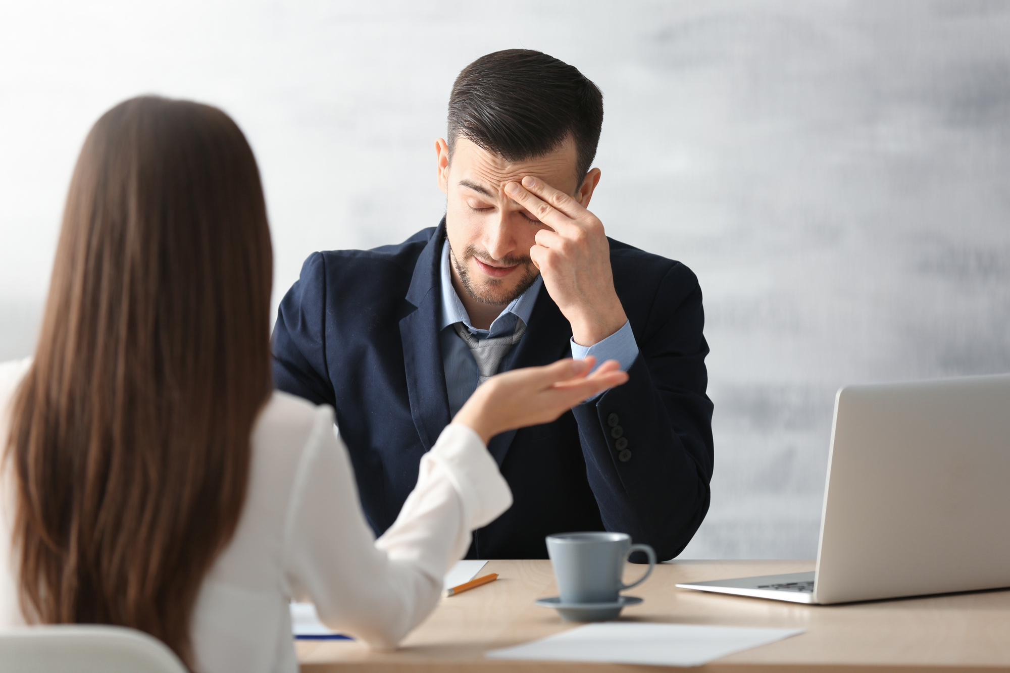 A man in a suit sits at a desk with a laptop, holding his forehead in frustration. A woman with long brown hair, seen from behind, gestures towards him as they have a conversation. A cup, papers, and pens are on the desk.