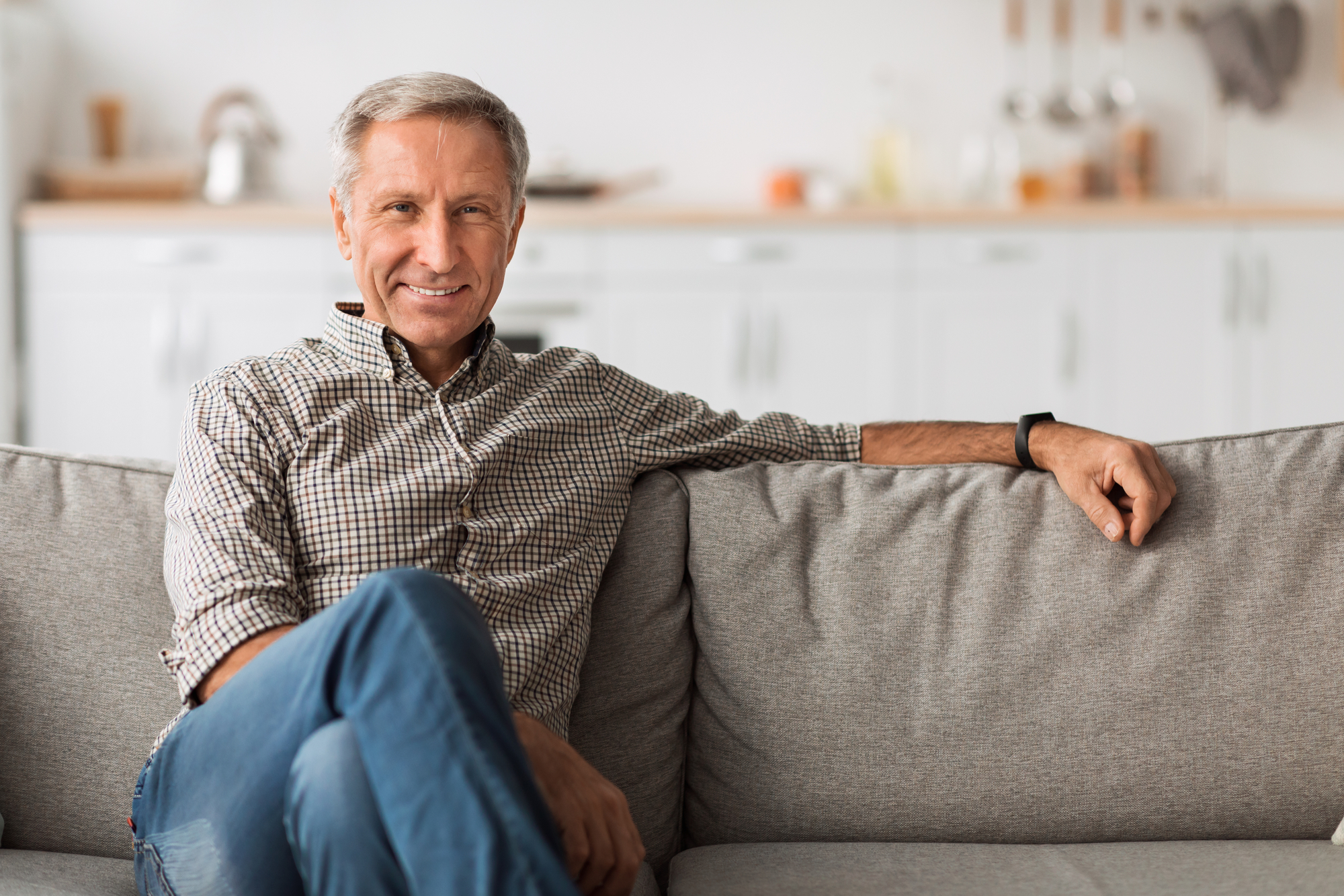 A smiling older man with short gray hair and wearing a plaid shirt and blue jeans sits on a gray sofa. He has one arm resting on the back of the sofa and the other on his knee. The background shows a bright kitchen with white cabinets.