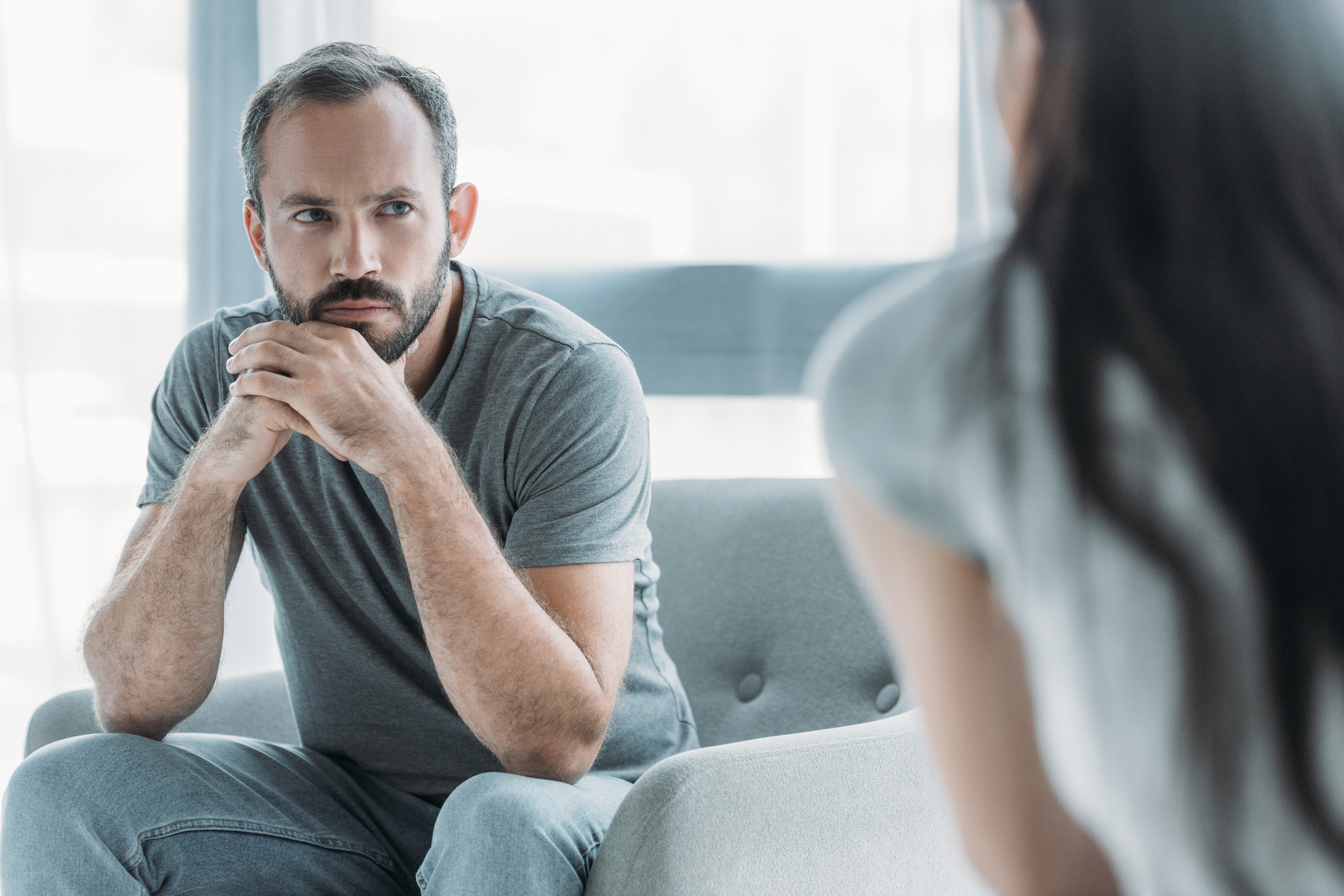 A man with a beard and short hair sits on a couch with his hands clasped under his chin, looking intently at someone who is partially visible in the foreground. He is wearing a gray t-shirt and jeans, and the background is softly lit and blurred.